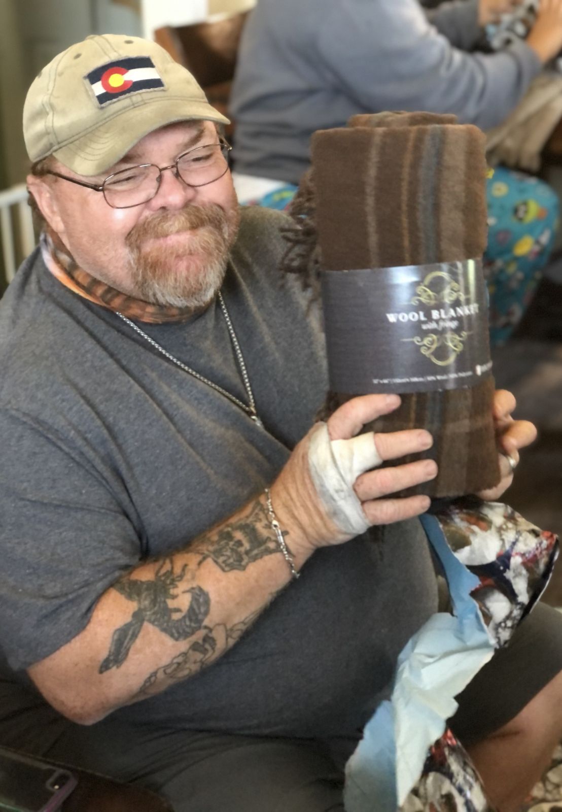 Neil seated holding a packaged blanket, tattoos visible on his arms