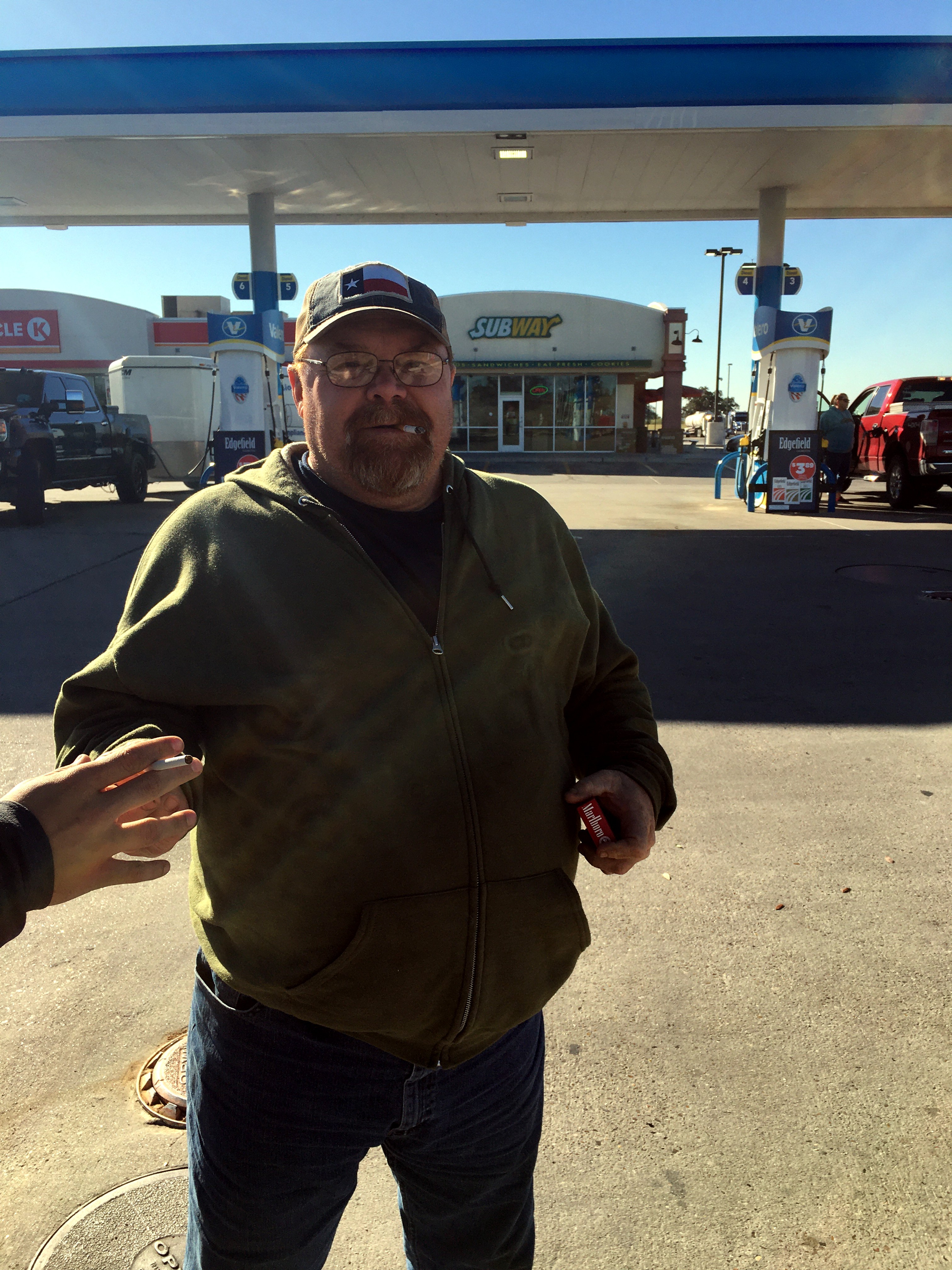 Neil at a gas station in a green hoodie and Texas flag cap